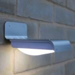 Lampe solaire LED murale en aluminium allumée, fixée sur un mur extérieur en briques, éclairage blanc intense.