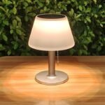 Lampe solaire LED à poser, abat-jour blanc et pied métal, éclairage chaleureux sur table en bois extérieur.