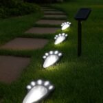 Chemin de jardin éclairé par des lampes solaires en forme d'empreintes de patte, intégrées dans l'herbe entre des dalles, avec un panneau solaire en arrière-plan et une végétation luxuriante