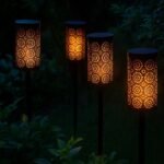 Quatre lampes solaires LED à effet flamme vacillante éclairant un jardin verdoyant au crépuscule, avec des motifs ajourés projetant une lumière chaleureuse dans la végétation.
