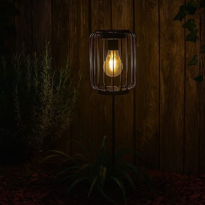 Lampe solaire LED blanc chaud en métal avec piquet, éclairant un jardin de nuit près d’une clôture en bois.