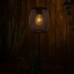 lampe-solaire-metal-jardin-nuit.jpg" alt="Lampe solaire LED blanc chaud en métal noir maillé, à piquer dans le jardin, éclairage décoratif IP44 de nuit devant une palissade en bois