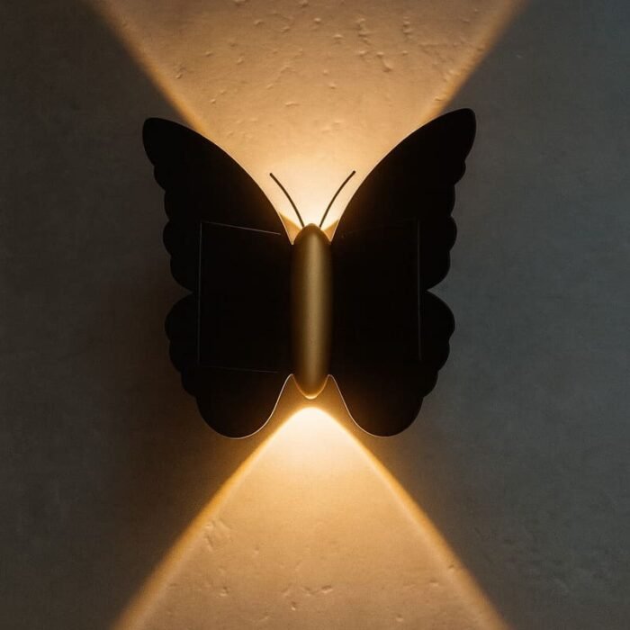 Applique murale solaire en forme de papillon noir et or allumée, diffusant une lumière chaude décorative en extérieur